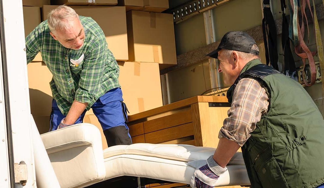 Zwei Männer heben in einem Umzugswagen voller Kisten ein Sofa an - Wohnen im Alter