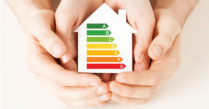 Ein Haus mit der Skala für die Energiewerte einer Immobilie wird von zwei Haenden festgehalten.