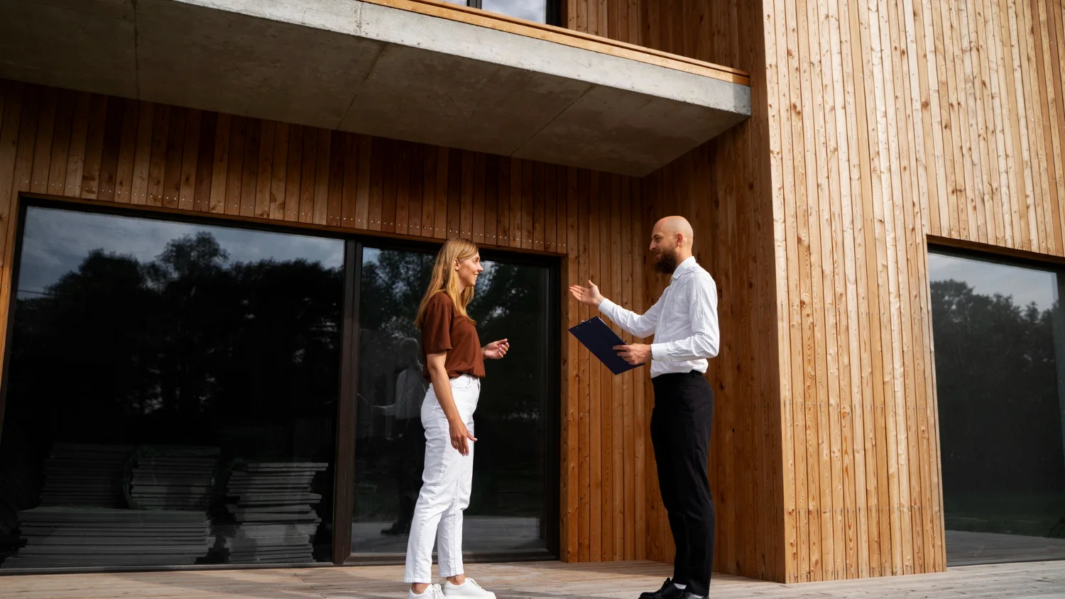 Immobilienmakler mit Unterlagen im Gespräch mit einer Eigentümerin vor einem modernen Einfamilienhaus mit Holzfassade